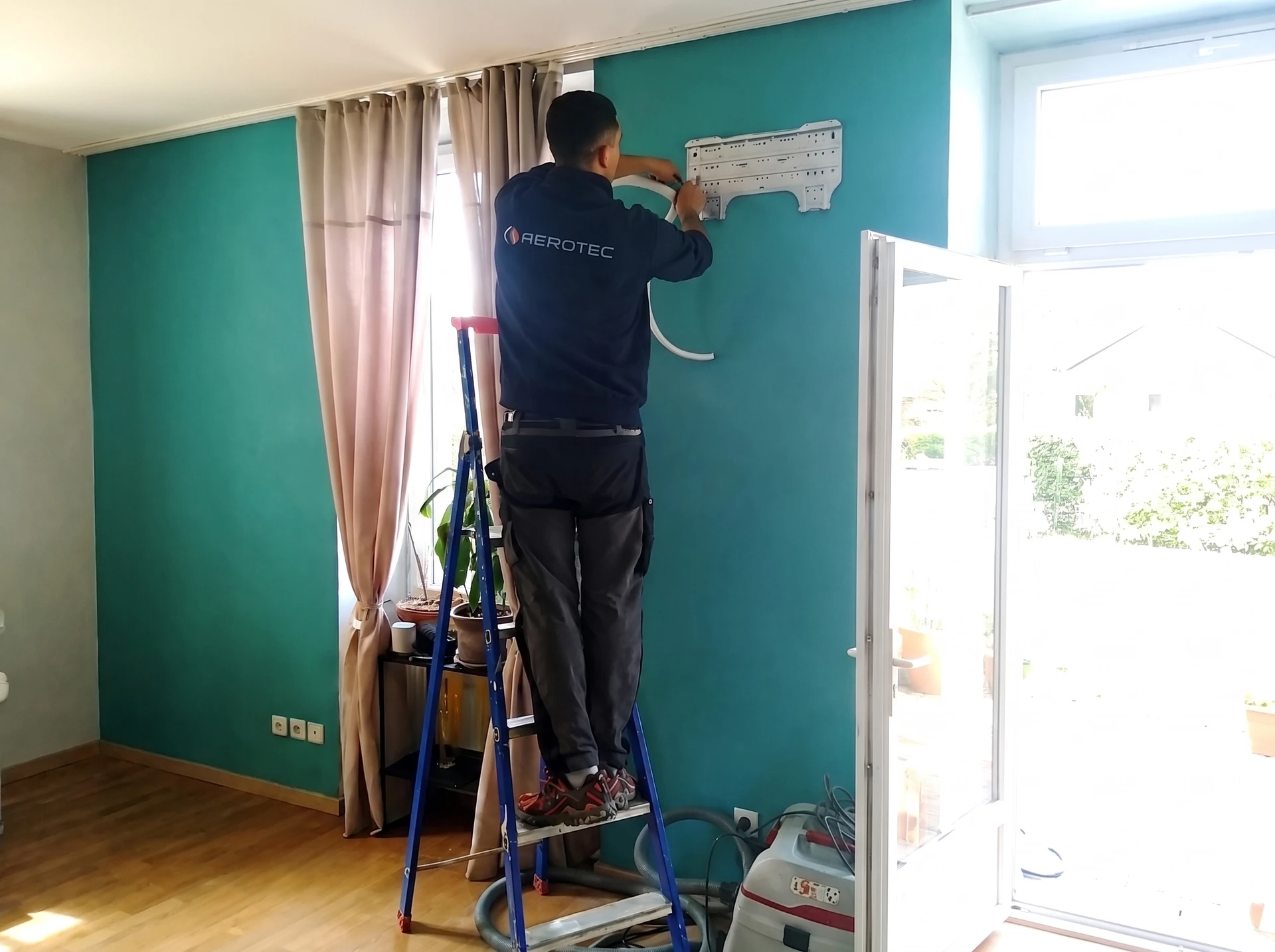 Technicien qualifié en combinaison de travail effectuant la maintenance d'une pompe à chaleur air-air, positionné devant l'unité intérieure murale du climatiseur réversible, en train de contrôler ou remplacer un élément interne tel qu'un filtre ou un composant électronique, dans un intérieur résidentiel moderne.